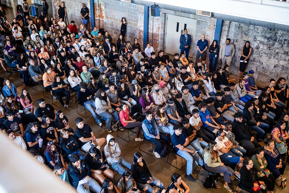 An overhead view of a large audience seated in rows during a Castro event at Metropolitan State University of Denver. Attendees are focused on the event, while some are using their phones. Several individuals stand along the back wall of the room.